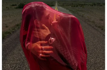 Een stelletje kust elkaar, bedekt met een rood doek, op een weg. Inez & Vinoodh, Think love, 2025